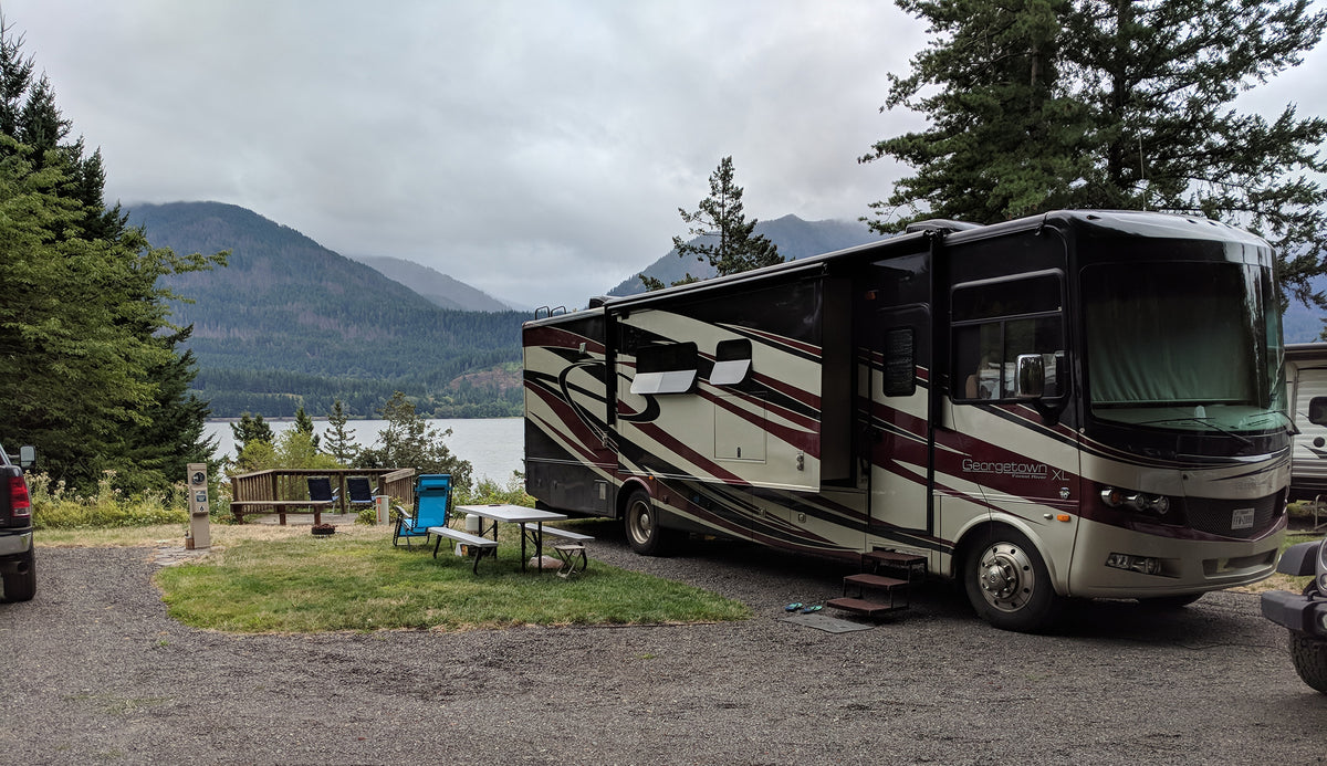 A large motorhome RV with its side door open, parked at a lakeside campsite with mountains and trees in the background