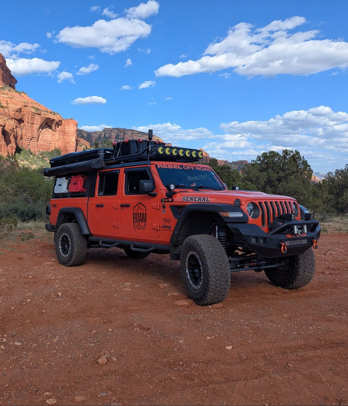 An orange off-road Jeep Wrangler parked on a red dirt road in a desert landscape with red rock formations, trees, and a blue sky with white clouds.