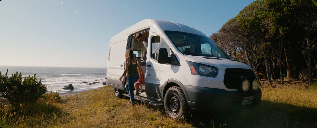 A white Ford Transit van parked outdoors on grass beside the ocean