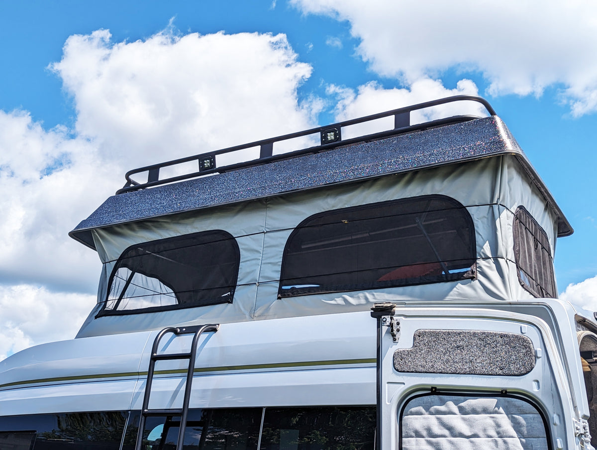Overland close-up of the upper section of a white motorhome or camper with a raised pop-top canvas roof, mesh window screens, a roof rack, and a ladder, photographed outdoors against a blue sky with clouds.