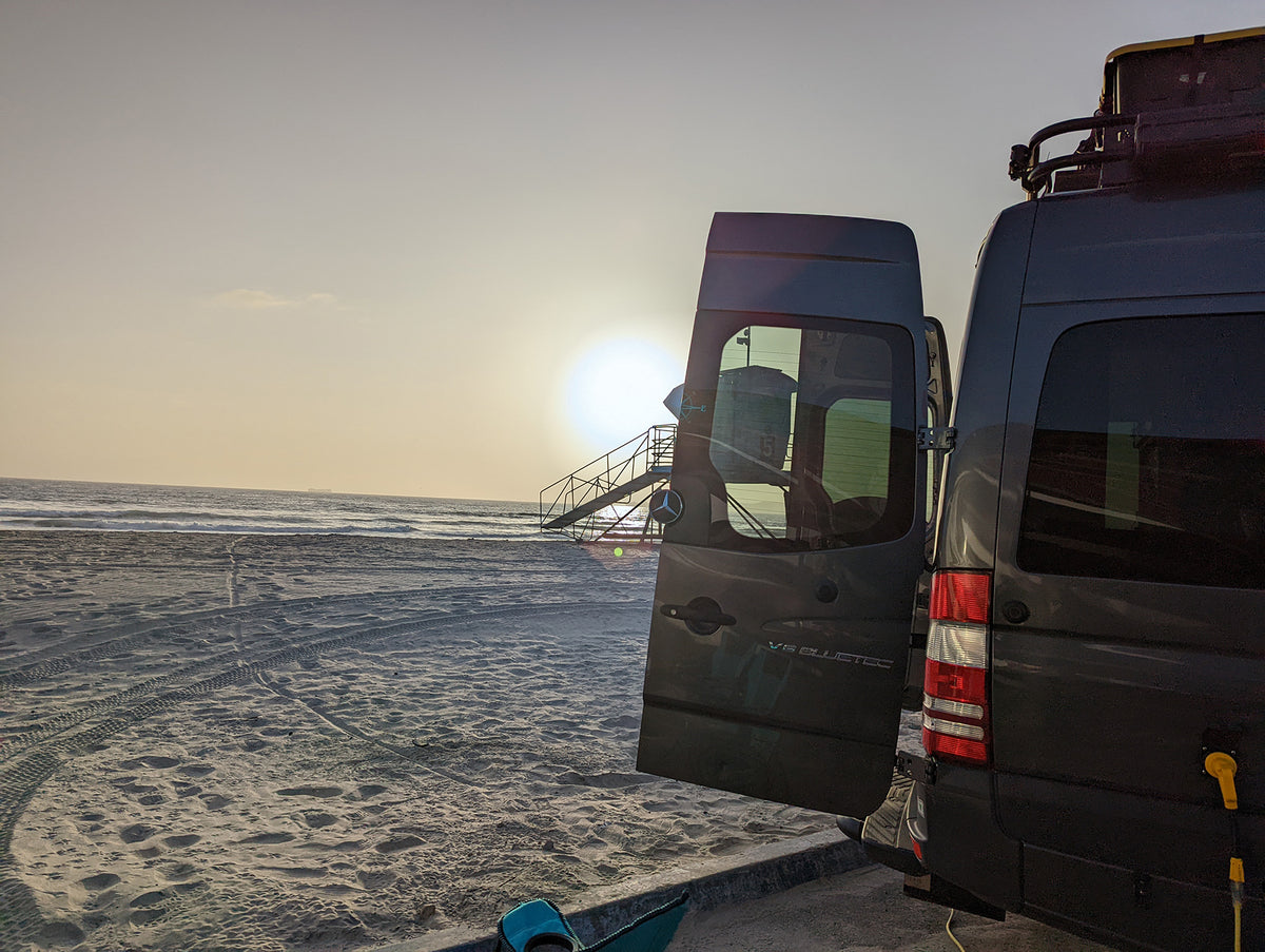 A dark gray camper van parked on a sandy beach at sunset, with the side door open and a staircase leading out of the vehicle. The background shows the ocean and a hazy sky.