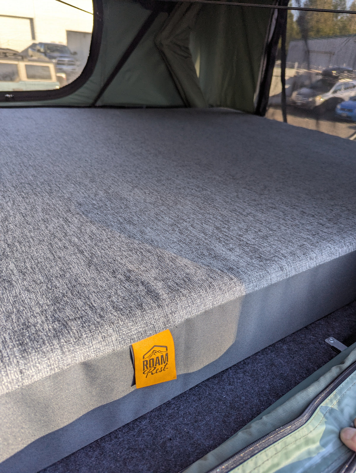 A close-up of a gray textured camping mattress or bed pad with a small yellow 'ROAD REEL' label, shown inside a tent with a window and outdoor parking lot visible in the background.