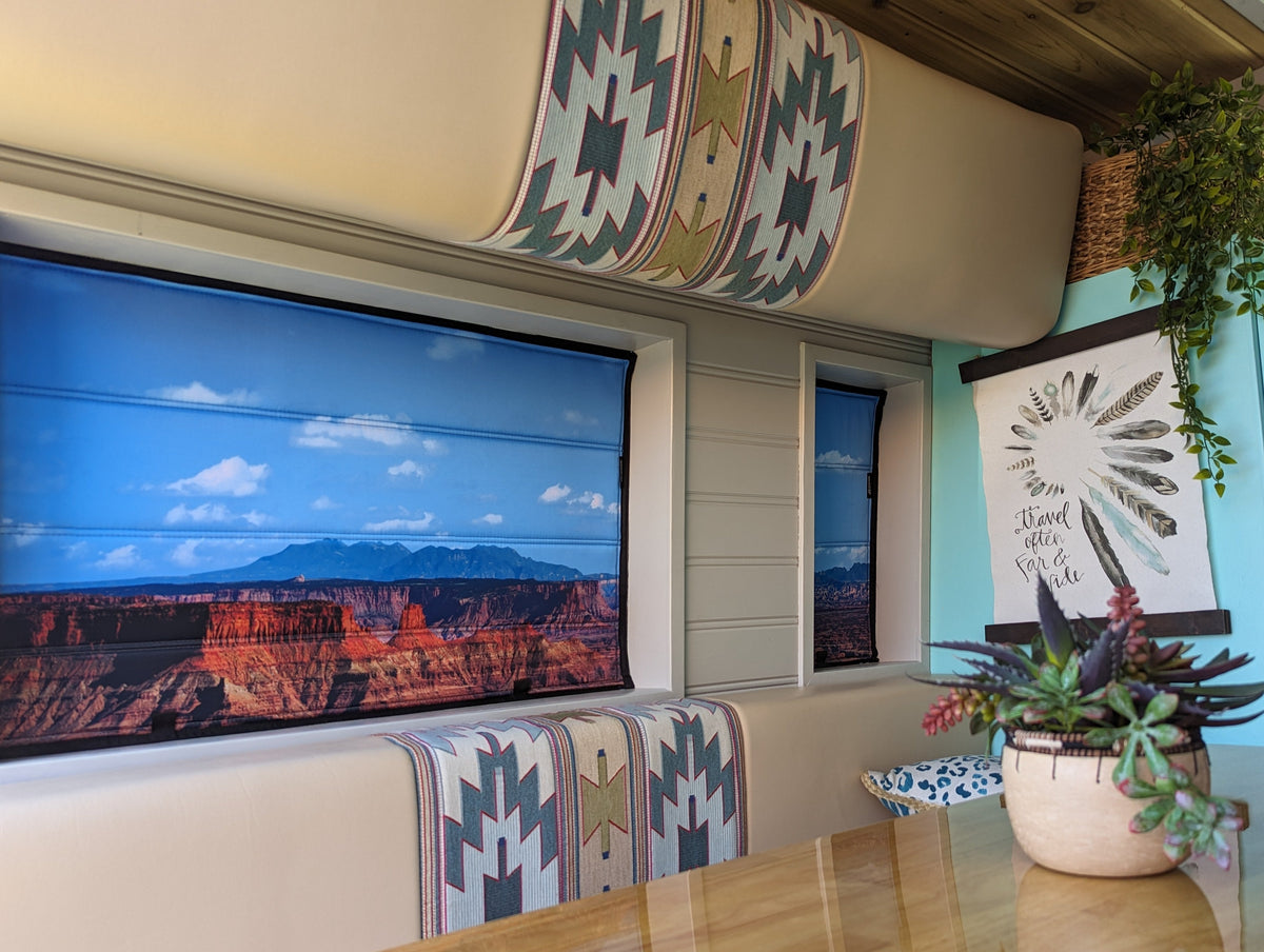 A rustic indoor dining area featuring a large wall-mounted screen displaying a canyon landscape, with matching smaller screens, patterned booth cushions, a wooden table, and a potted succulent.