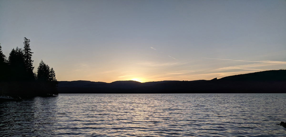 A scenic landscape photograph of a lake at sunset, featuring dark silhouetted hills, pine trees on the left, and the sun low on the horizon reflecting over the water.