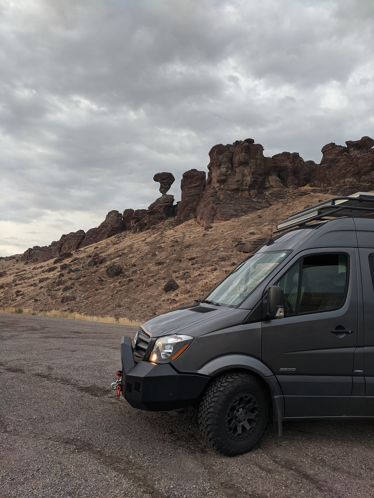 A dark gray Mercedes-Benz Sprinter van parked on a gravel road with a rocky desert hillside and cloudy sky in the background.