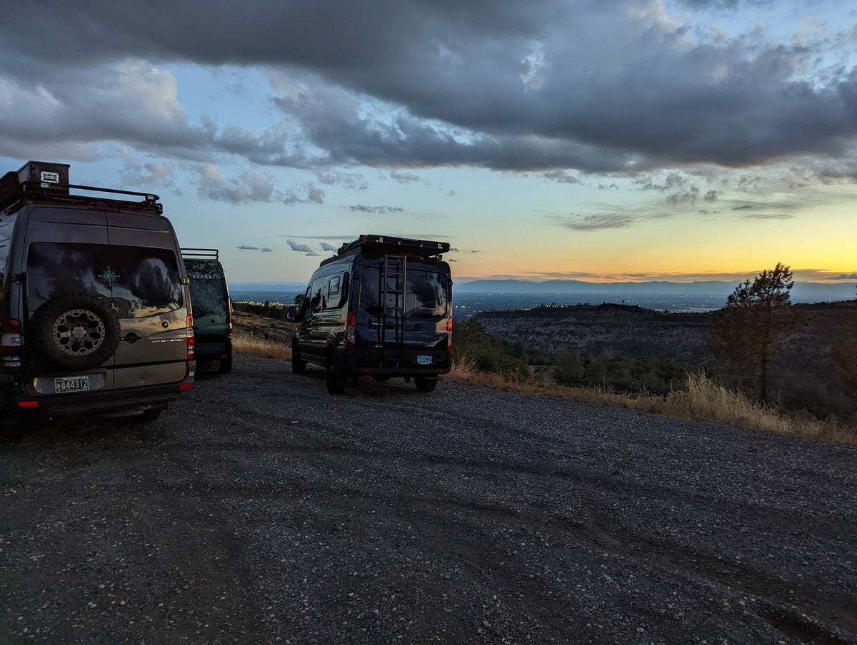 Three travel vans parked on a gravel road at dusk overlooking a mountain landscape under a dramatic cloudy sky.