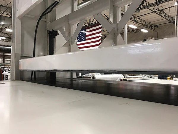 A large white rectangular panel or beam resting horizontally in a warehouse or factory setting, with industrial metal beams, overhead lighting, and an American flag visible in the background.