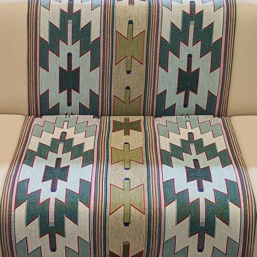 A close-up of a woven textile with a Southwestern geometric pattern, draped over a beige upholstered chair. The pattern features dark green, beige, and light blue zigzags and crosses with red and blue border stripes.